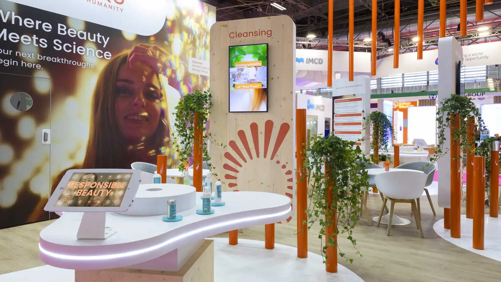Un stand d'exposition moderne et esthétique avec une ambiance naturelle. Des plantes grimpantes entourent des poteaux orange, et un écran digital affiche des visuels sur un panneau en bois clair. Le design du stand présente un mélange de matériaux naturels et de technologie, avec des produits de beauté exposés sur une table blanche aux formes arrondies. Une grande image de fond montre une personne souriante, avec des lumières brillantes pour attirer l'attention des visiteurs.